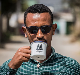 Den Becher in der rechten Hand haltend trinkt ein Mann mit Sonnenbrille aus dem Moyee Coffee Emaille Cup