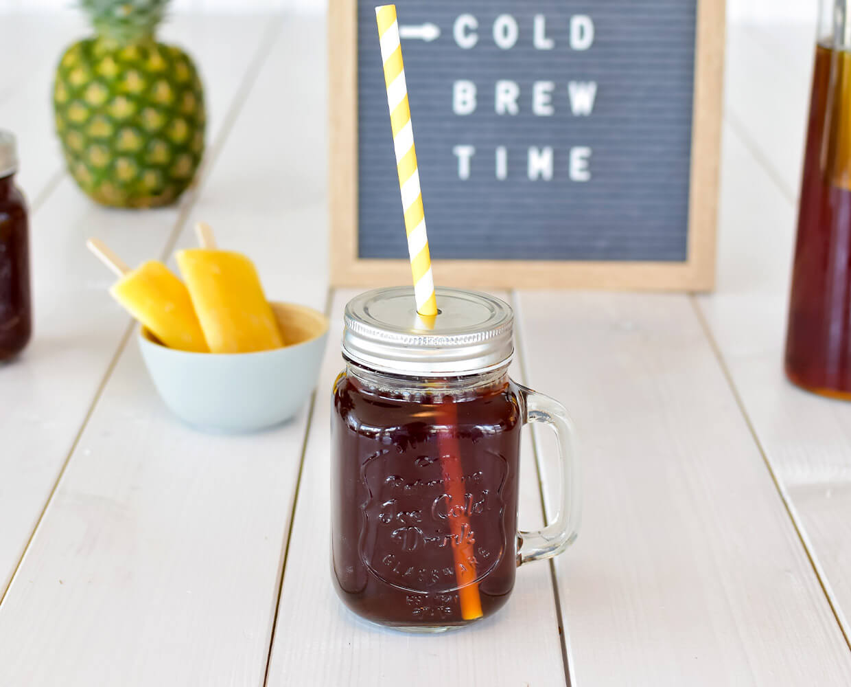 Vorne steht ein Glas mit Cold Brew Kaffee und Strohhalm und dahinter Eis am Stiel, eine Ananas und ein Schild mit dem Schiftzug Cold Brew Time