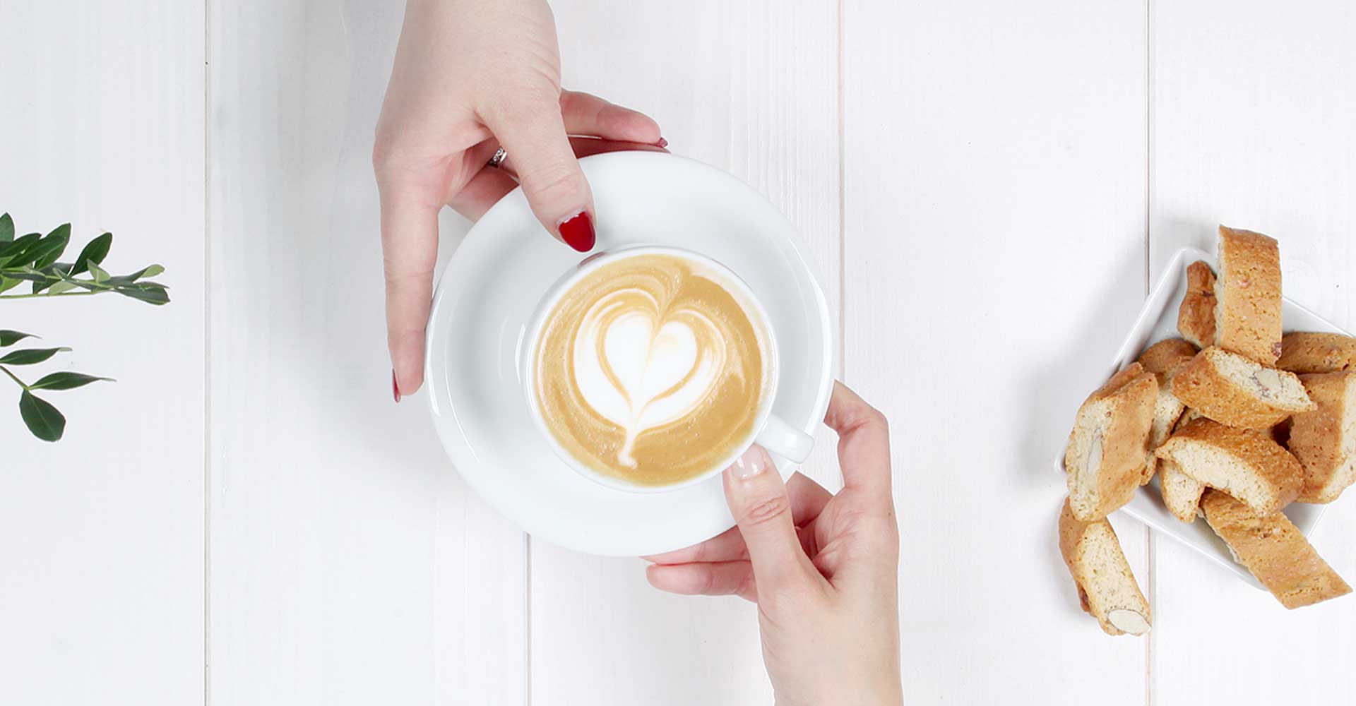 Eine Hand überreicht einer anderen einen Cappuccino mit Latte Art, dazu gibt es Cantuccini