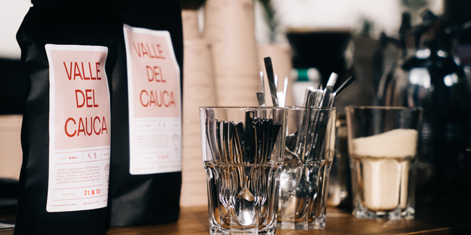 Zwei Valle Del Cauca Kaffeetüten stehen neben Gläsern mit Kaffeebesteck