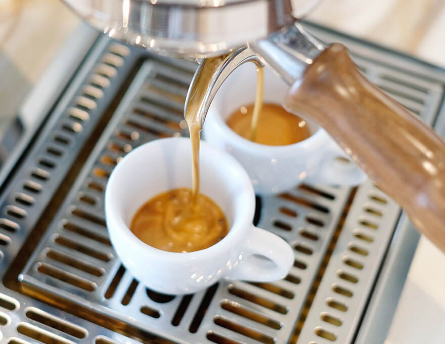 Zwei weiße Espressotassen stehen auf einem Gitter und aus einem Siebträger mit Holzgriff läuft Espresso in die Tassen.